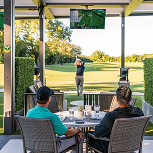 Members seated at tables watching a golfer practice on the range