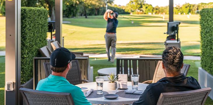 Guests seated on a patio watching a golfer hit balls at a driving range