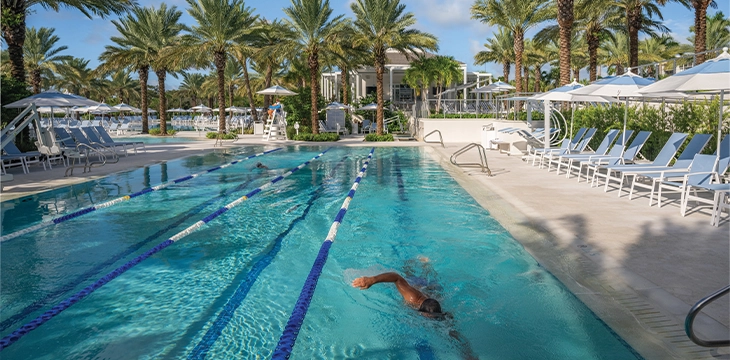 Outdoor lap pool with swimming lanes and a swimmer, surrounded by palm trees and lounge chairs.