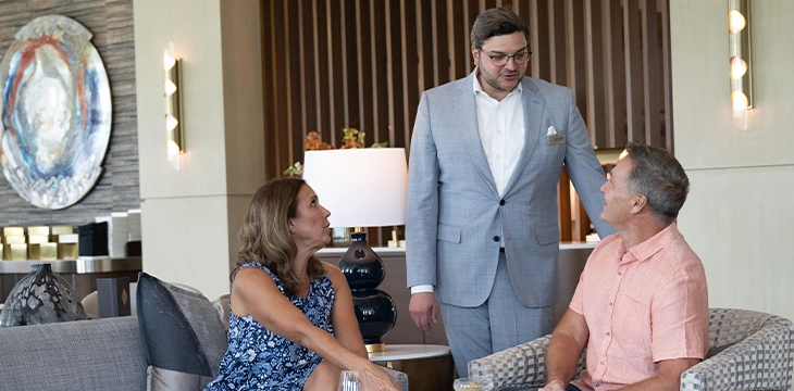 Hotel staff member in suit speaking with a seated couple in a lounge area with wine glasses on the table.
