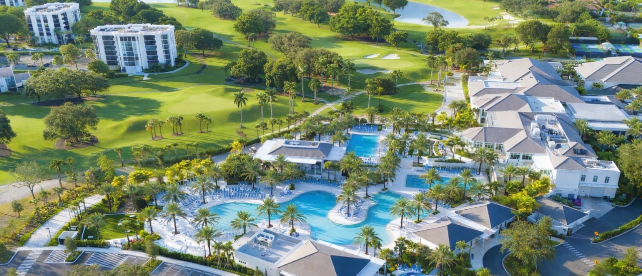 An aerial drone perspective shows a large resort complex with multiple interconnected swimming pools surrounded by palm trees and lounge areas. Nearby are manicured golf course fairways and greens with sand bunkers. Residential buildings and landscaped pathways border the pool complex.