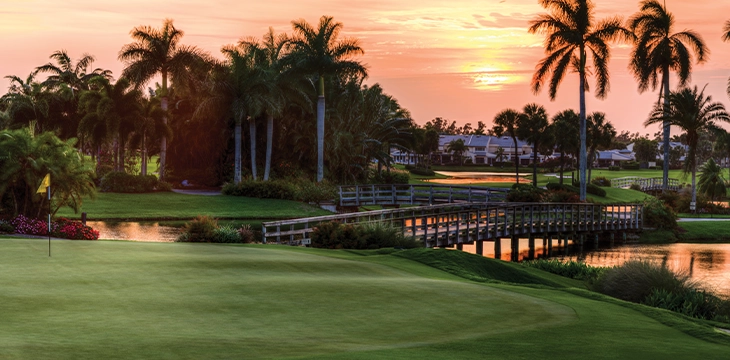 Golf course at sunset with palm trees and a bridge over water.