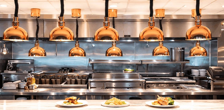 Open restaurant kitchen with copper heat lamps over plated dishes.