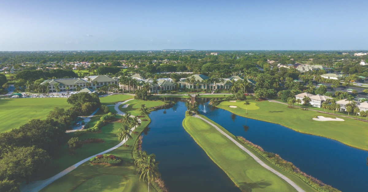Aerial view of a golf course with lakes, palm trees, paths, and clubhouse buildings under a clear sky.