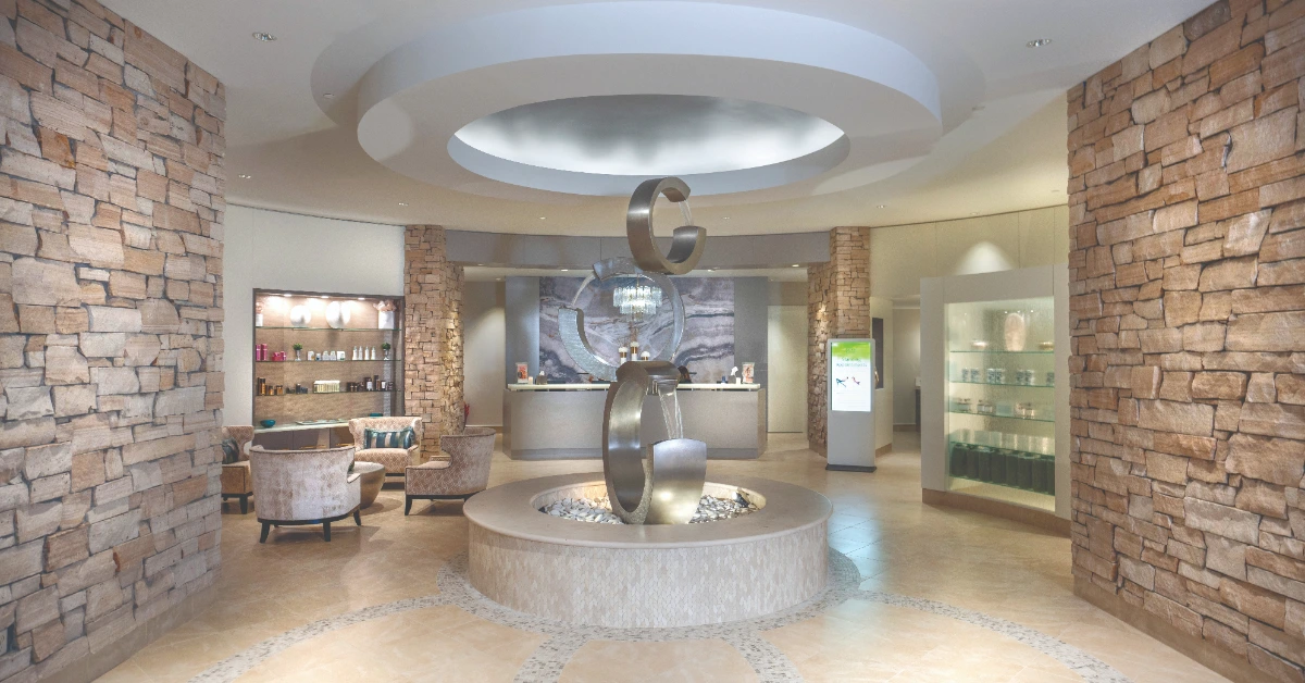 Bright spa lobby with stone columns, circular ceiling feature, reception desk, and abstract sculpture centerpiece.