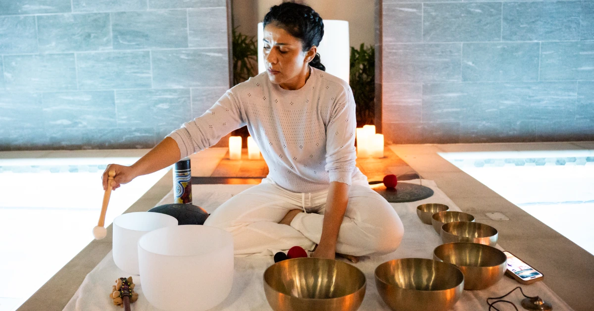 Woman in white plays crystal and brass singing bowls in a candlelit spa room beside illuminated pools.