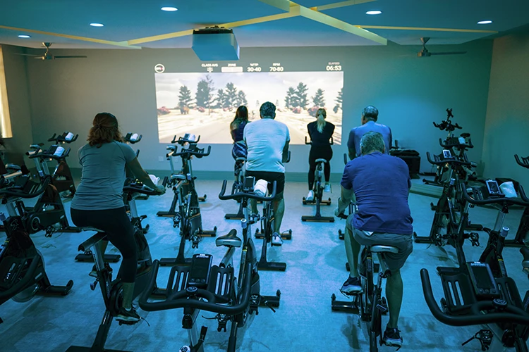 People riding stationary bikes in dark studio with screen