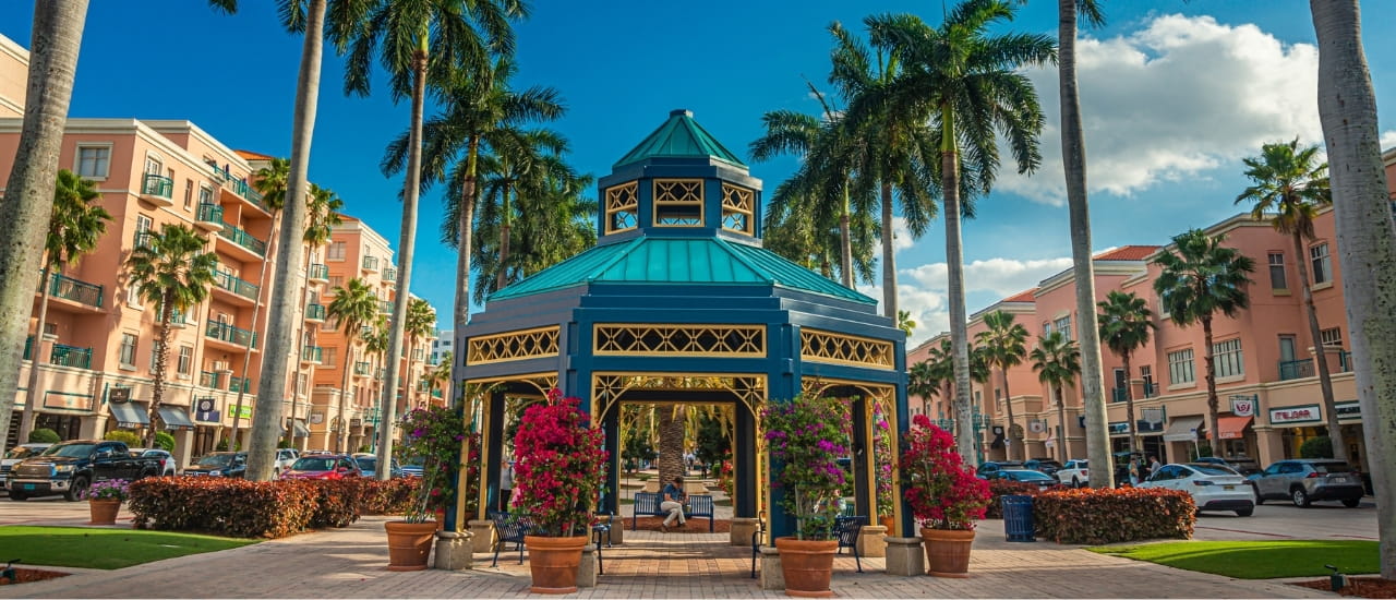 A vibrant town square gazebo surrounded by royal palm trees and colorful bougainvillea blooms in a sunny Florida coastal community.