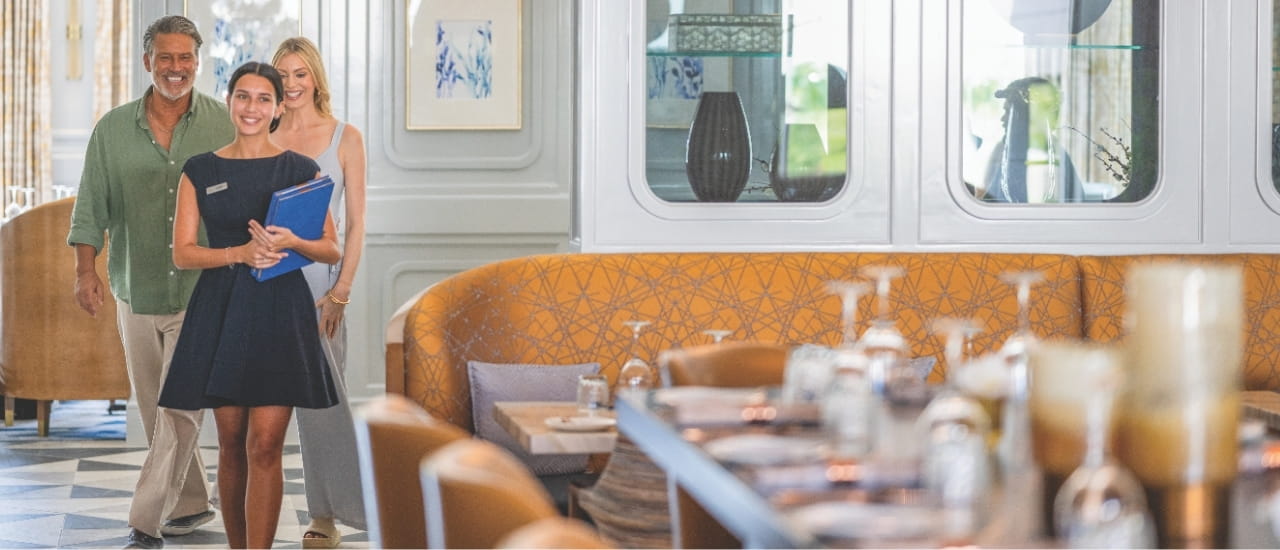 A smiling hostess welcomes guests into an elegant restaurant dining room featuring warm leather seating and sophisticated interior design.
