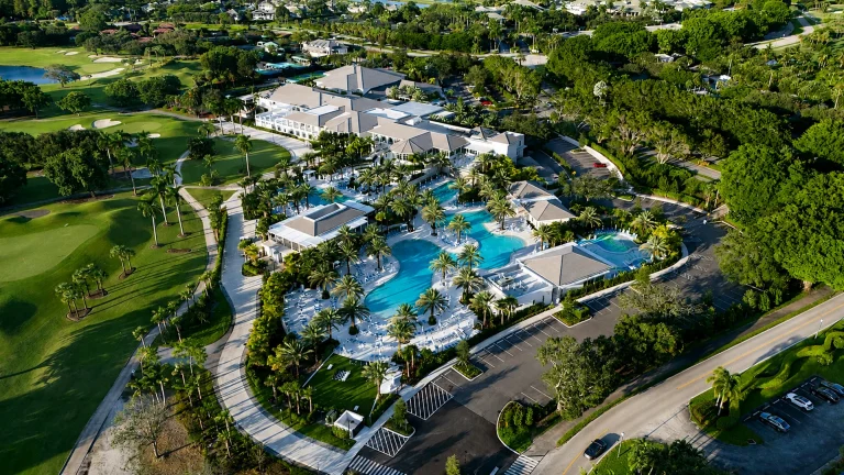 Aerial daytime view of a large clubhouse building surrounded by pools, palm trees, and a golf course.