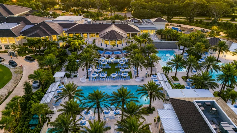 Aerial twilight view of a lit-up clubhouse building beside a glowing swimming pool with blue umbrellas.