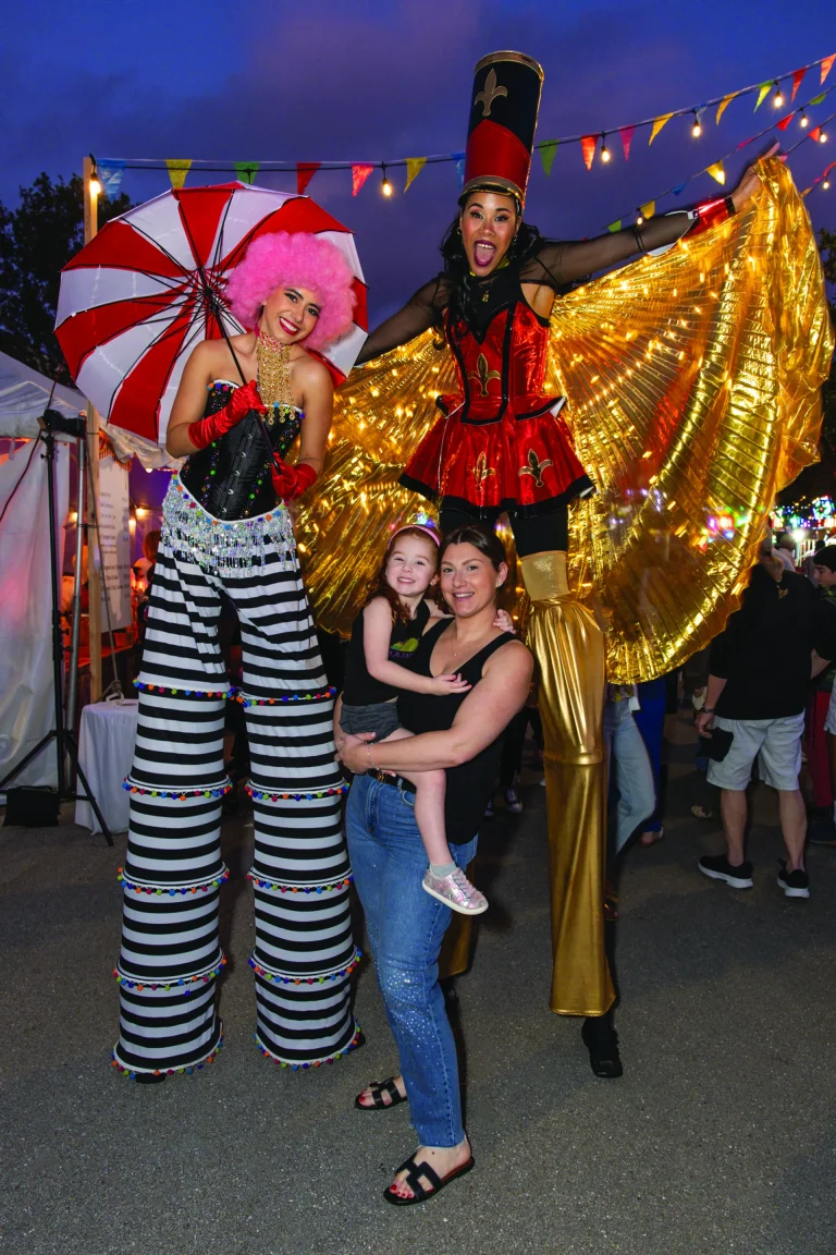Two costumed stilt performers posing with a mother and child at a Boca West fair-themed event.