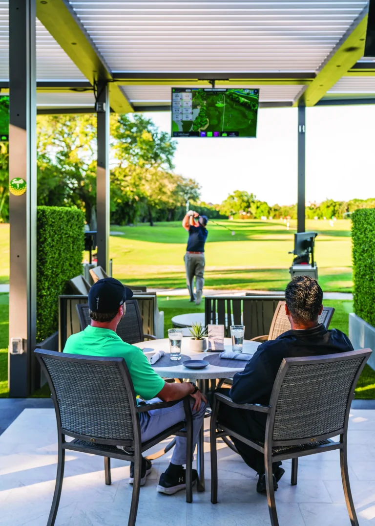 Members seated at a table under a covered golf bay overlooking the Boca West driving range.