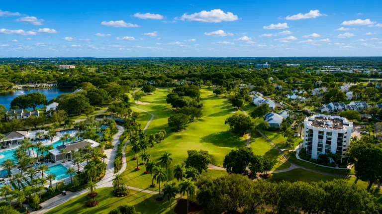 Aerial view of a golf course fairway surrounded by palm trees, low-rise condos, a lake, and a swimming pool complex.