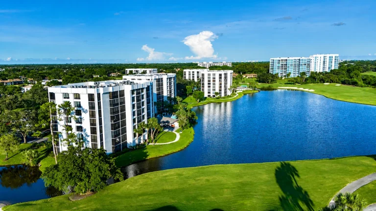 Several white mid-rise condo buildings along the edge of a blue lake, with a golf course in the background.